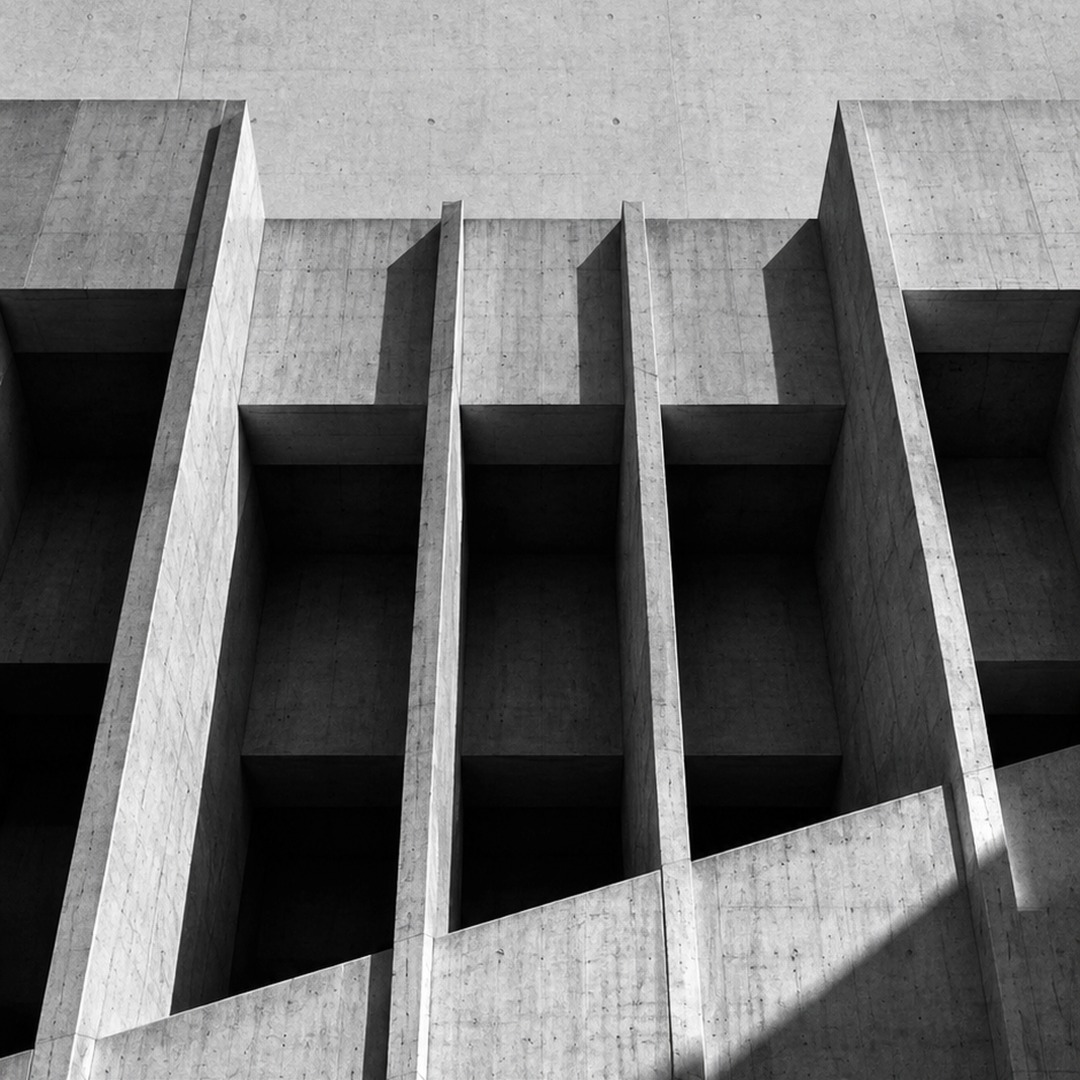 Black-and-white photograph of a concrete 
									building with repeating vertical columns and angular shadows